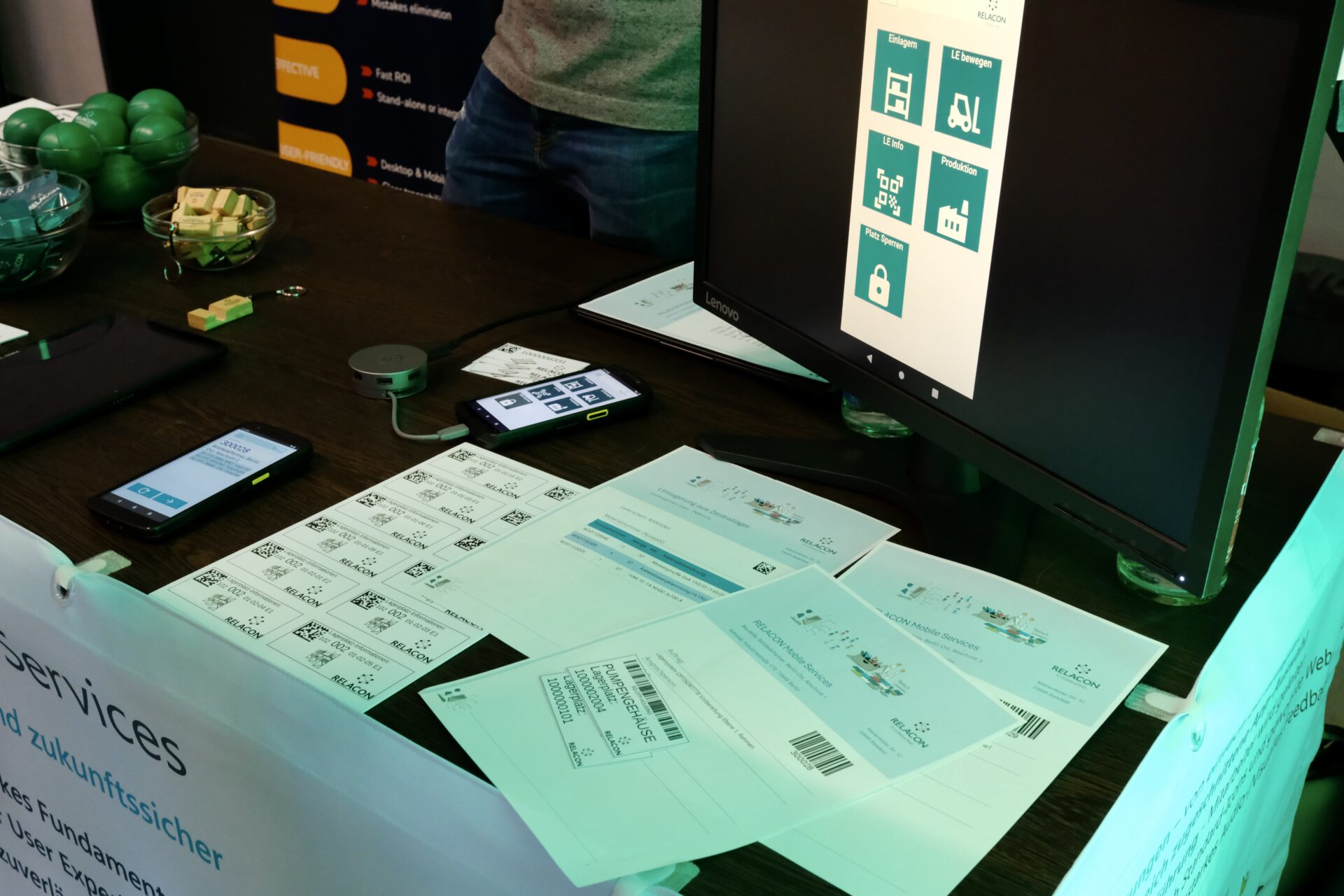 RELACON Mobile Services Demo-Setup – Monitor zeigt intuitive App-Oberfläche mit Prozess-Icons, mobile Endgeräte und Labelprint-Beispiele auf dem Tisch
