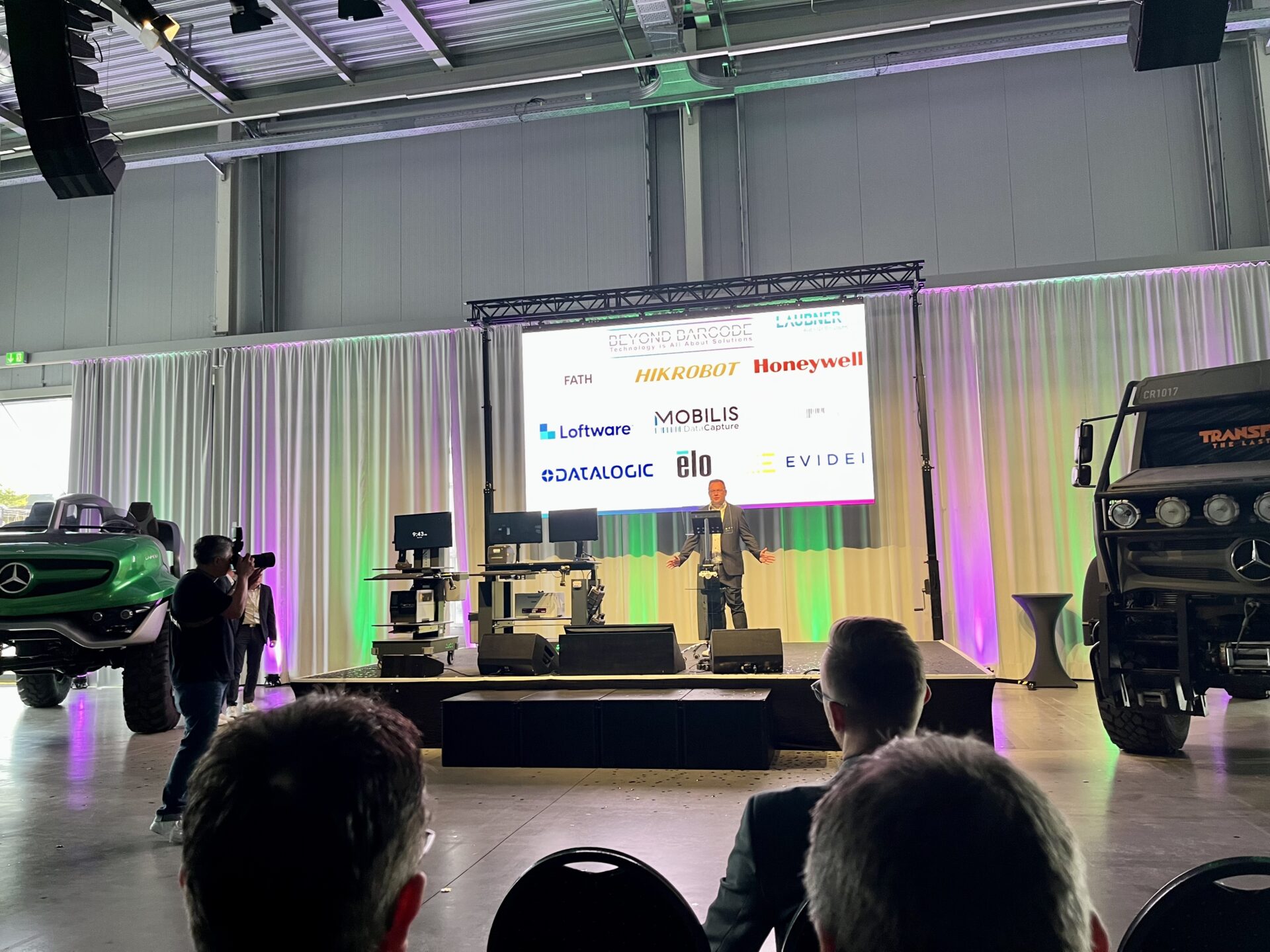 BEYOND BARCODE Messebühne im Hangar 10 des Technikmuseums Speyer – Redner vor Partnerleinwand mit Logos von Honeywell, HIKROBOT, Datalogic, MOBILIS und weiteren, flankiert von historischen Fahrzeugen