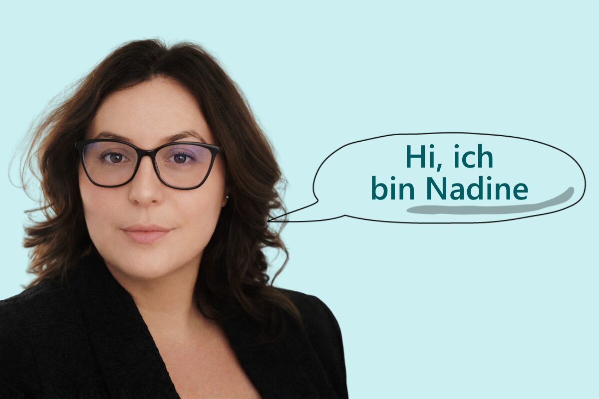 Foto von Nadine, einer Frau mit langen, dunkelbraunen Locken und schwarzer Brille, in schwarzem Blazer vor hellblauem Hintergrund. Neben ihr eine Sprechblase mit dem Text: „Hi, ich bin Nadine".