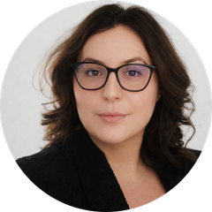 Porträtfoto von Nadine, einer Frau mit langen, dunkelbraunen Locken und schwarzer Brille, in schwarzem Blazer vor hellem Hintergrund.